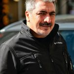 Smiling mature man posing outdoors in a black jacket, captured in natural daylight.